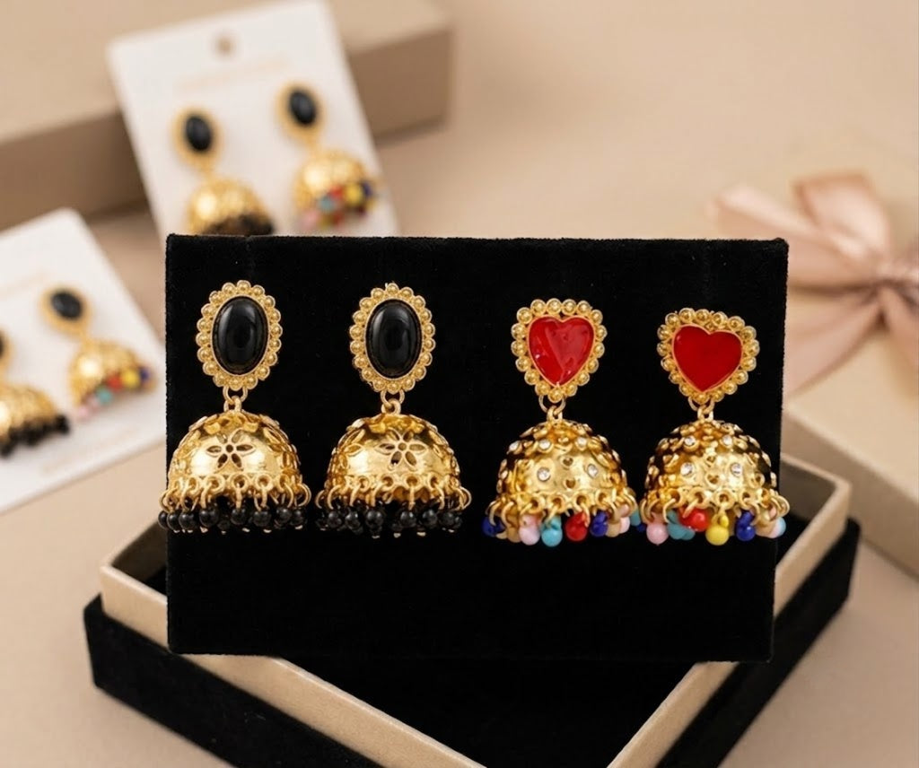 Four pairs of gold-toned jhumka earrings displayed on a black velvet jewelry box. The pairs feature black and red oval and heart-shaped stones, with one pair adorned with small multicolor bead drops.